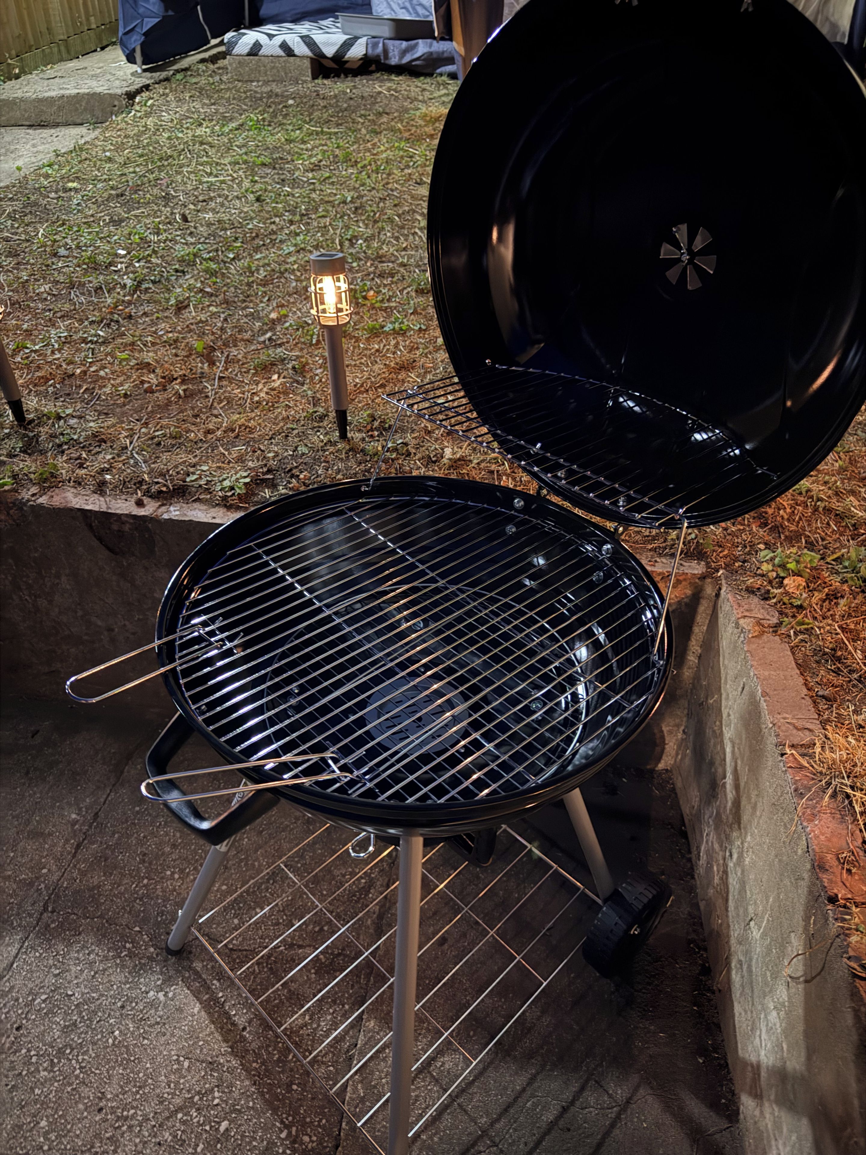 Ein schwarzer Grill mit Drahtrost, geöffnetem Deckel und einer kleinen Lampe am Boden.
