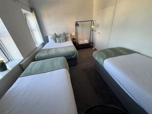Una habitación de hotel con tres camas, una ventana, un radiador y una mesita de noche con lámpara.