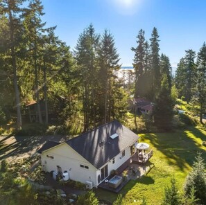 A single-story house with a dark roof, surrounded by dense forest and a clear sky. - Whidbey Island Oasis with Hot Tub and Fire pit (Freeland)