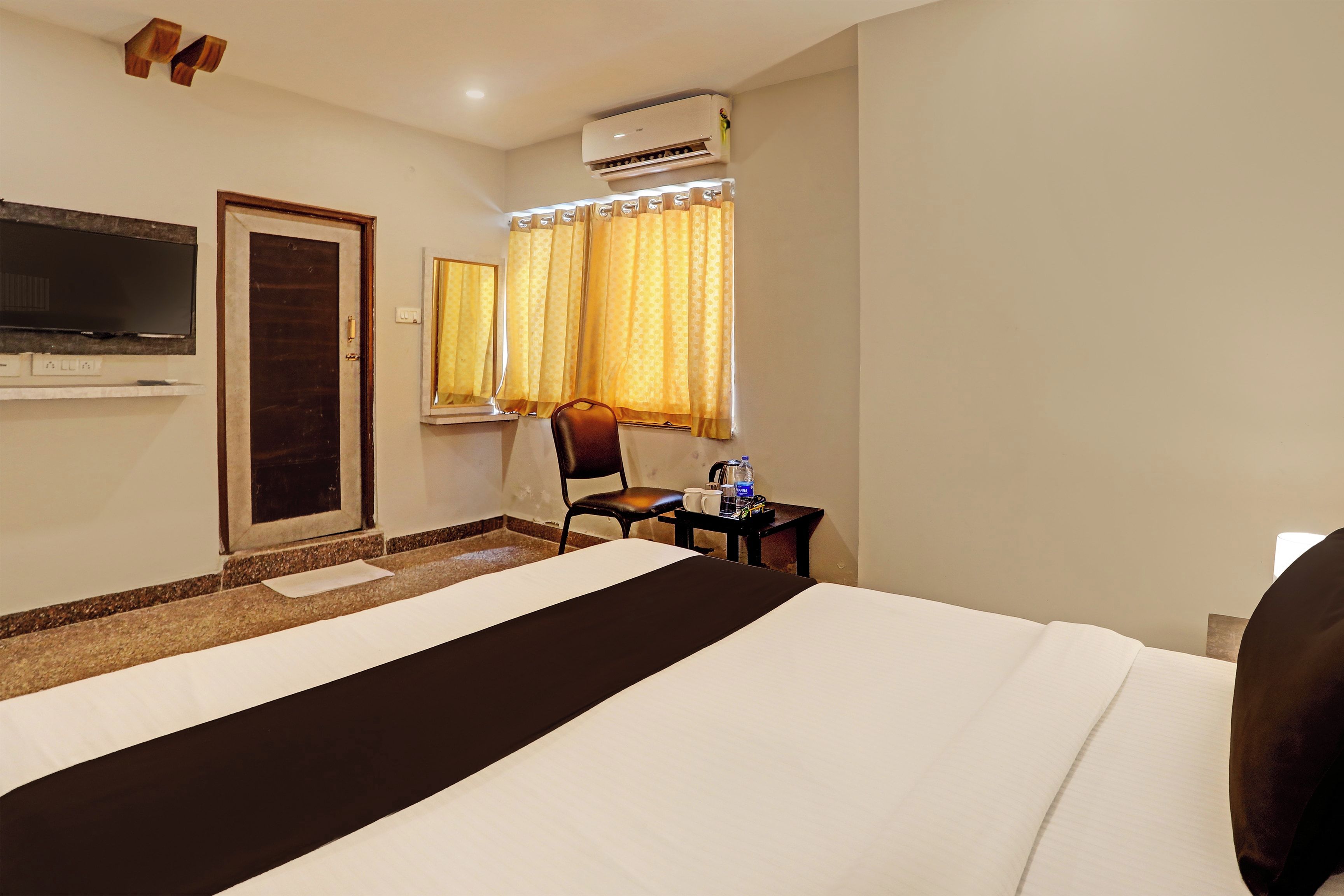 Habitación de hotel con una cama, televisión montada en la pared, una silla, una mesita con una botella de agua y una taza, y una ventana con cortinas.