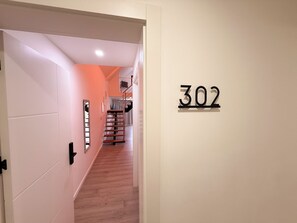 Een gang met een houten trap, een deur met het nummer 302, en een lamp aan het plafond.