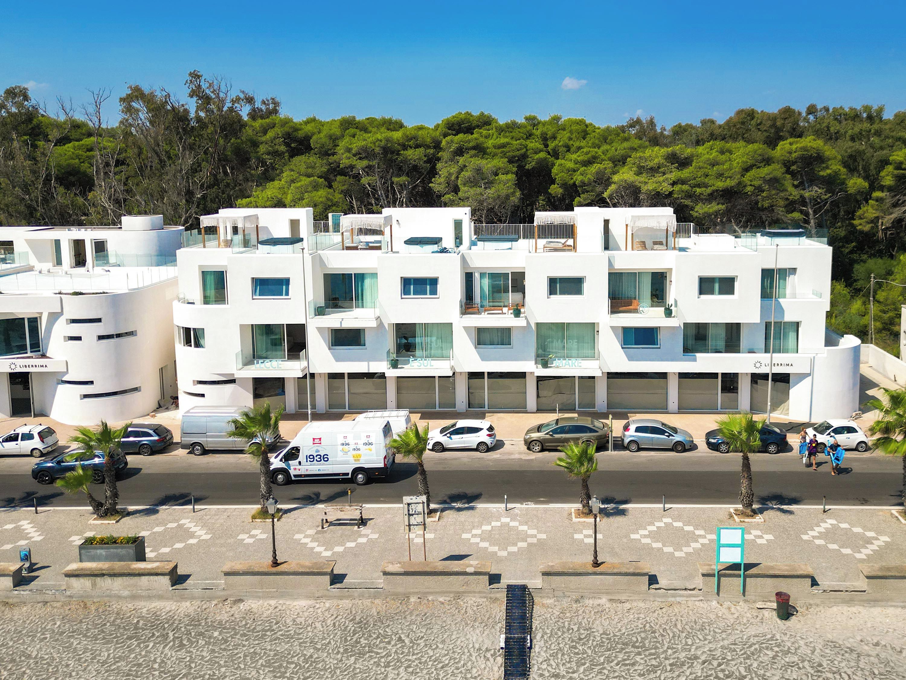 A modern multi-story hotel with a white facade, multiple balconies, and a parking area with several cars.