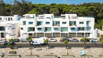 A modern multi-story hotel with a white facade, multiple balconies, and a parking area with several cars.