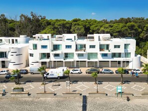 Un hotel moderno de varios pisos con fachada blanca, múltiples balcones y un estacionamiento con varios coches.