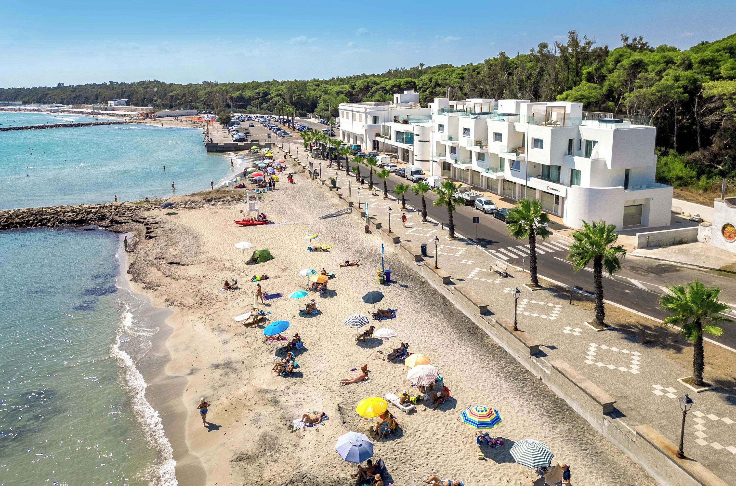 A beachside hotel with a row of rooms along the shore, a sandy beach with umbrellas, and a clear view of the sea.