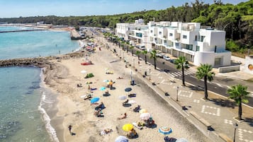 A beachside hotel with a row of rooms along the shore, a sandy beach with umbrellas, and a clear view of the sea.