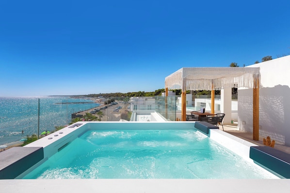 Una piscina infinita en la azotea con vistas despejadas al mar y un edificio blanco con una pérgola de madera.