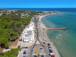 Vista aérea de una zona costera con un camino que lleva a una playa, coches aparcados y un edificio grande con techo plano.