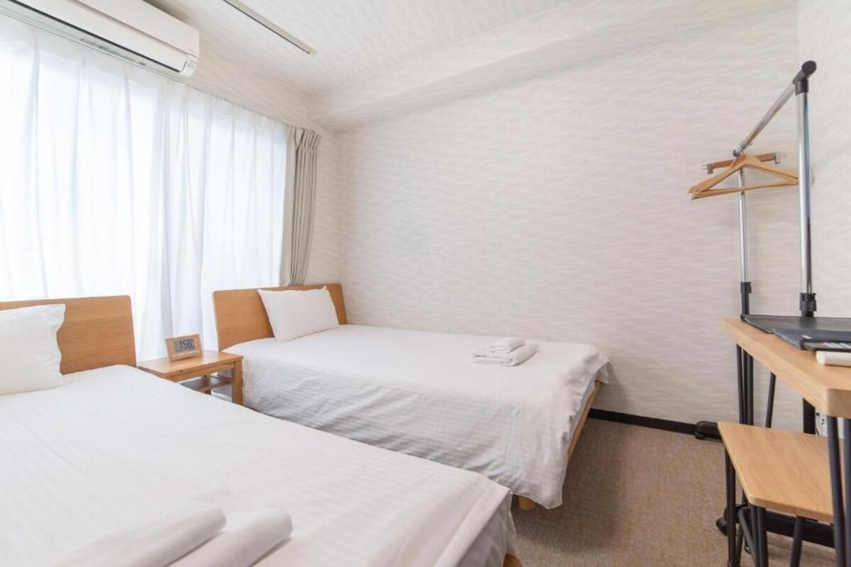 Et hotelværelse med to senge, et skrivebord, en stol, et knagered og et vindue med gardiner.