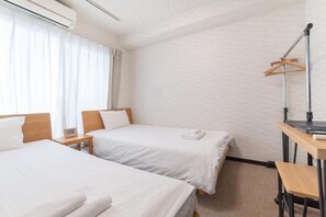 Et hotelværelse med to senge, et skrivebord, en stol, et knagered og et vindue med gardiner.