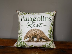 Un cojín cuadrado con una ilustración de pangolín y el texto 'Pangolin's Rest' sobre una pared lisa.