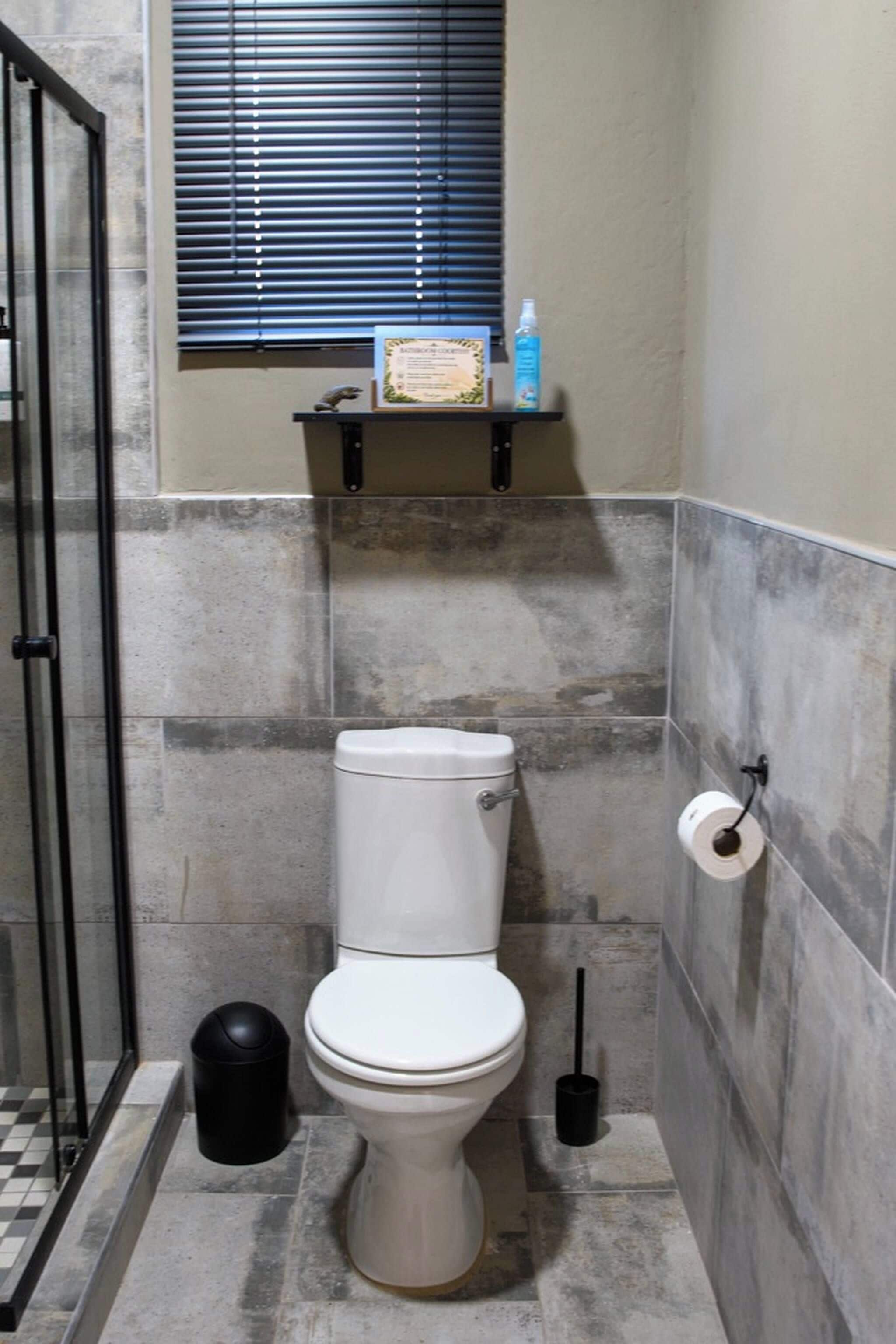 A bathroom with a toilet, a trash bin, a toilet paper holder, and a window with blinds.