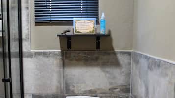 A bathroom with a toilet, a trash bin, a toilet paper holder, and a window with blinds.