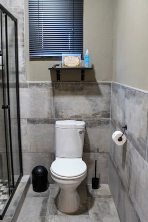 A bathroom with a toilet, a trash bin, a toilet paper holder, and a window with blinds.