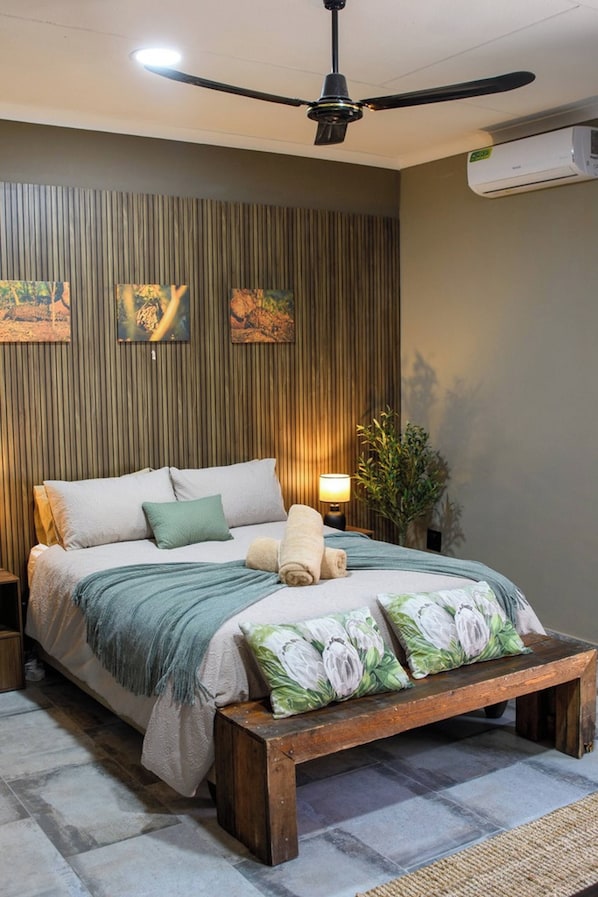 A bedroom with a bed, pillows, a bedside table, a lamp, a bench, a ceiling fan, and wall-mounted artwork.