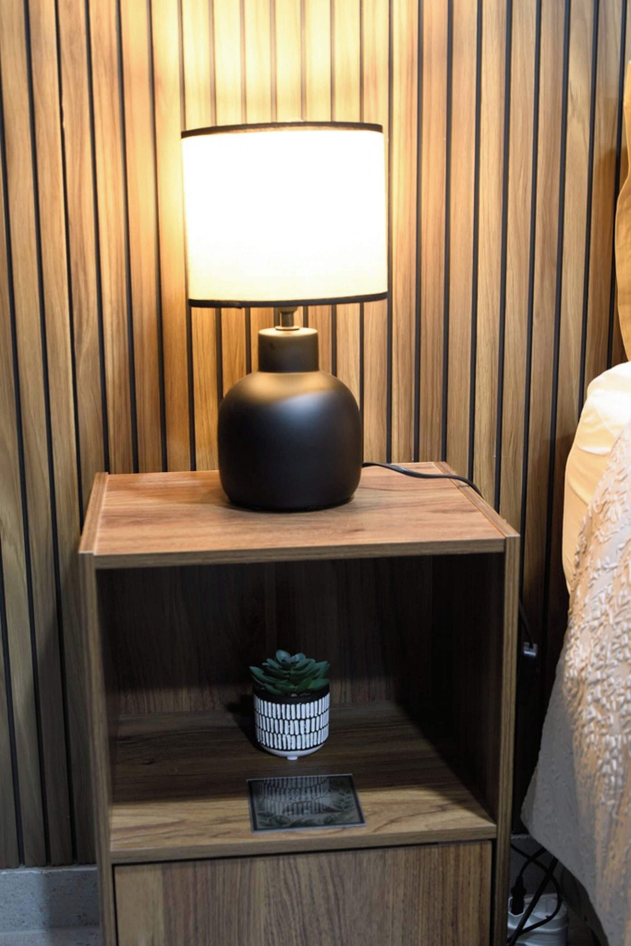 A bedside table with a lamp, a small shelf with a plant, and a framed picture.