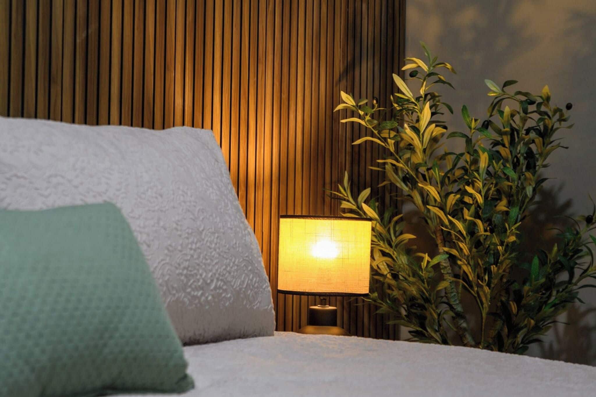 Un lit avec des oreillers, une lampe de chevet et une plante en pot, dans une pièce aux murs lambrissés.