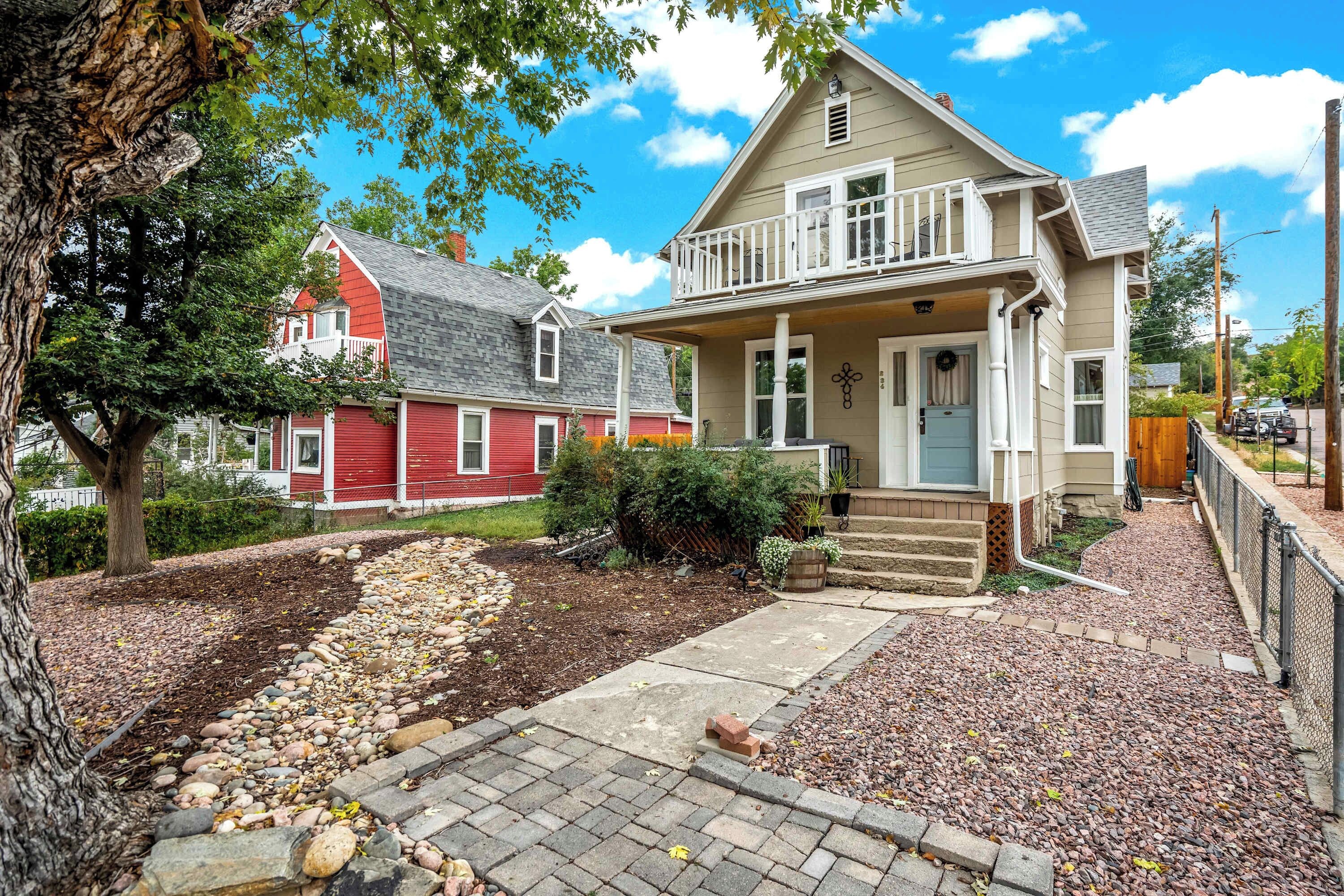 A two-story house with a front porch, a small garden with a stone pathway, and a red house in the background.