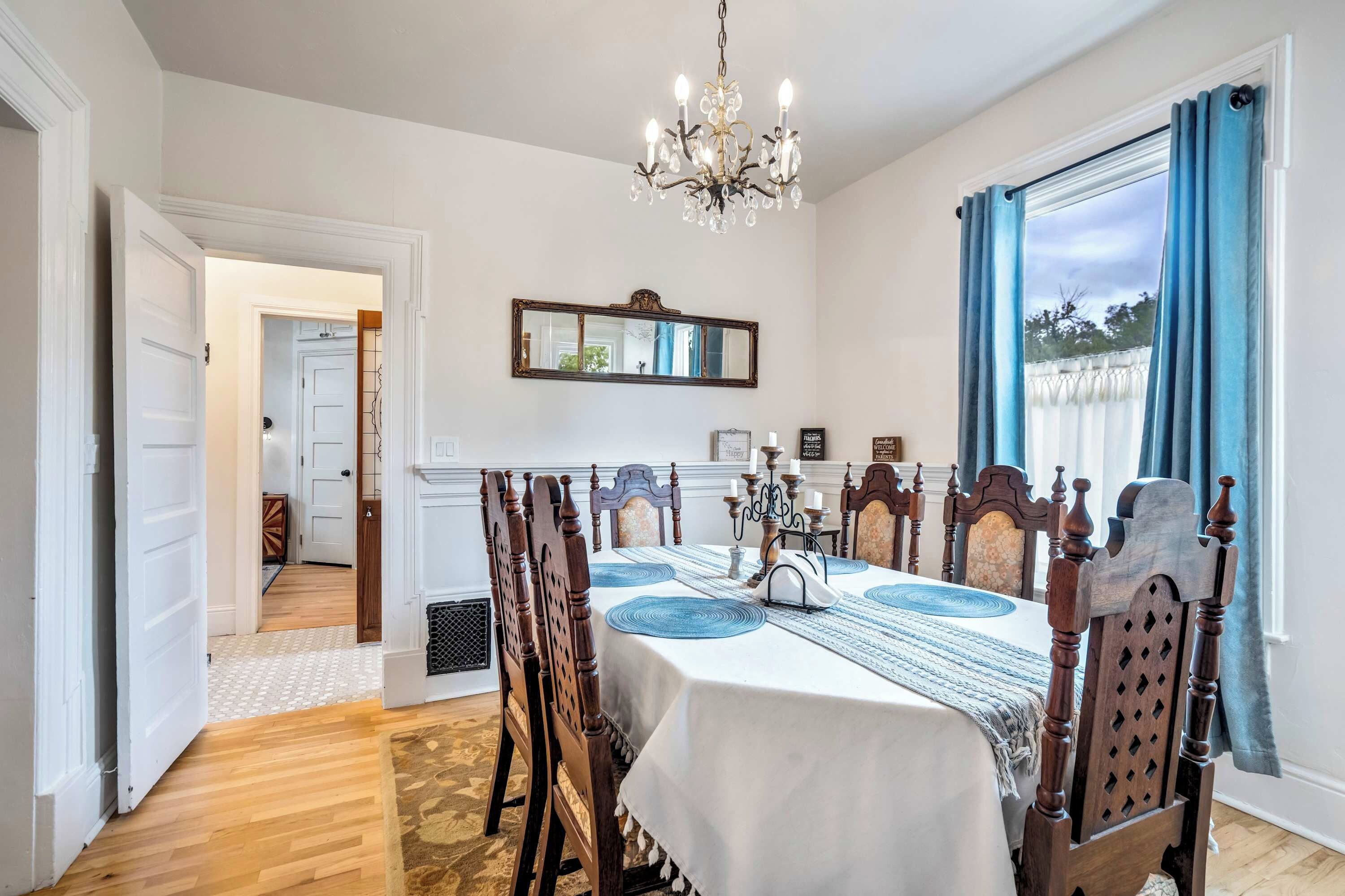 A dining room with a chandelier, wooden chairs, a table with a white tablecloth, and a view through a window with blue curtains.