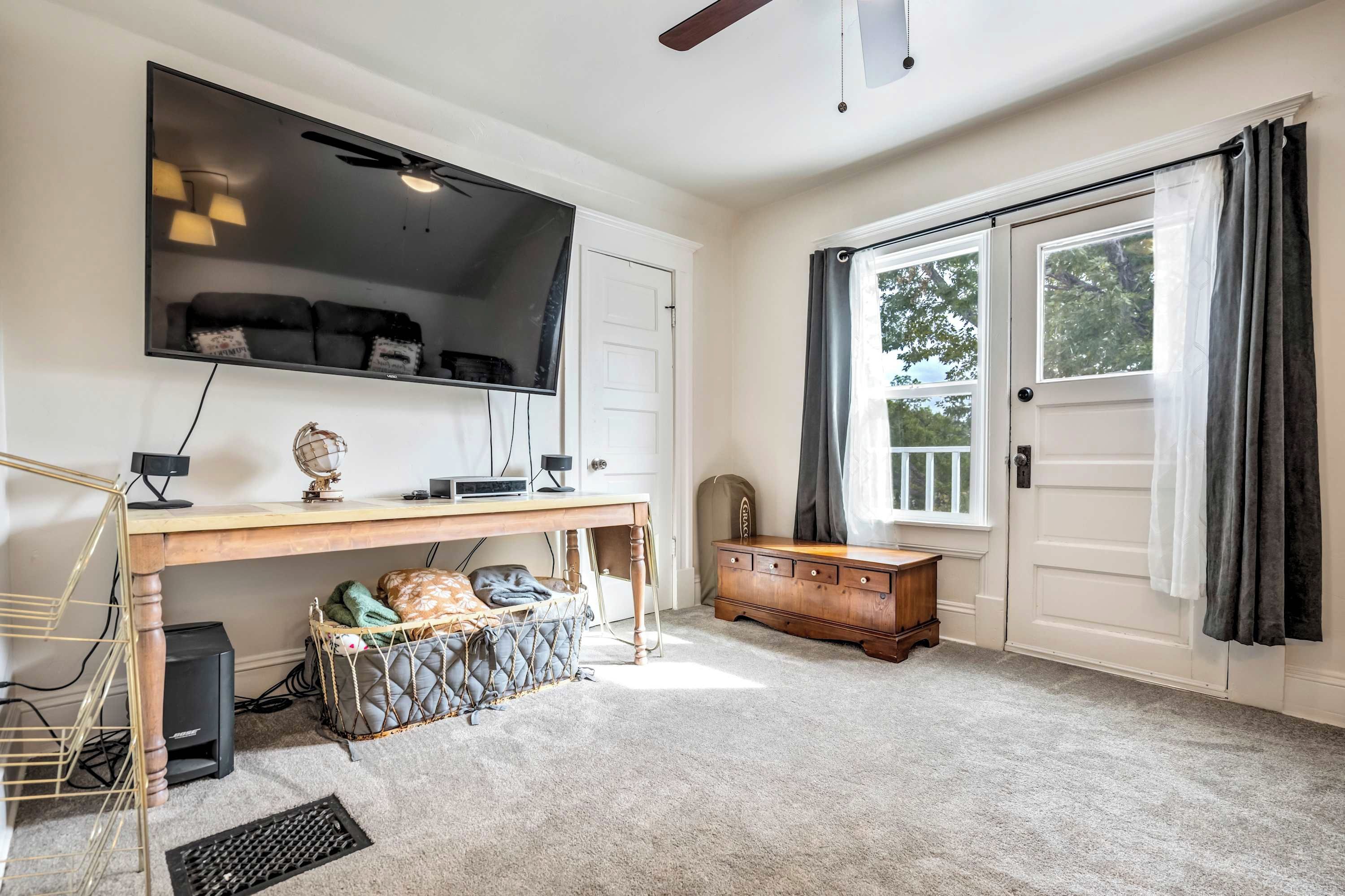A room with a large flat-screen TV mounted on the wall, a wooden desk with a computer and a globe, a cushioned bench with a basket, a wooden cabinet, and a sliding glass door leading to an outdoor area.