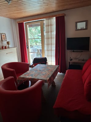 Ein Zimmer mit Holzboden, einem an der Wand montierten Fernseher, einem Tisch mit einer blumigen Tischdecke, roten Stühlen und einem Sofa.