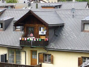 A traditional chalet with a wooden balcony, a satellite dish, and a flag. - Cozy 2.5- (3)-room apartment, large kitchen, rooms facing the garden, south-facing, no traffic (Scuol)