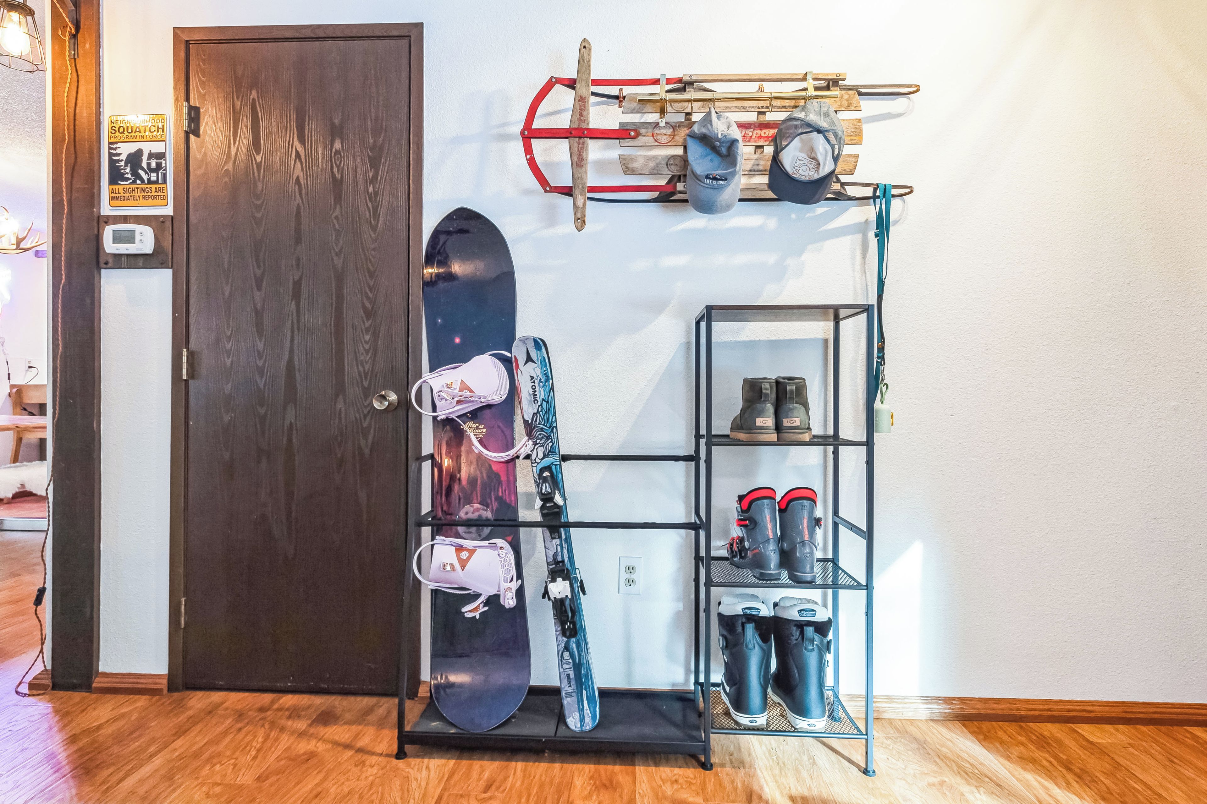 A room with a wooden door, a wall-mounted rack holding pots and a snowboard, and a shelf with boots and a helmet.
