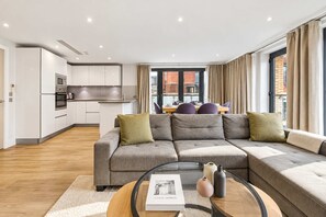 Un salon moderne avec un canapé d’angle, une table basse en verre et une vue sur la ville grâce à de grandes fenêtres.