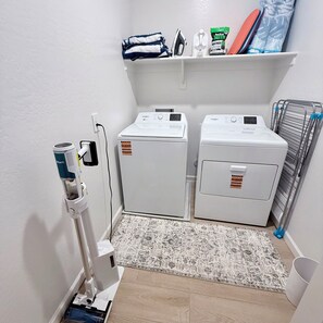 A laundry room with two front-loading washing machines, a wall-mounted ironing board, and a shelf with folded towels and cleaning supplies. - South Mountain 4 bedroom luxury retreat (Phoenix)