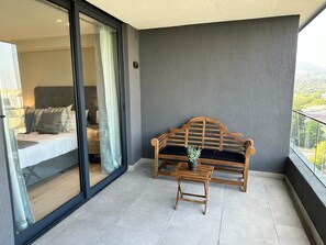 A balcony with a wooden bench and a small table, a sliding glass door leading to a bedroom, and a view of greenery. - Newlands Peak Penthouse (Cape Town)