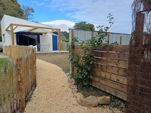Un sendero de grava conduce a un portón de madera, un edificio blanco con una puerta de garaje azul y una valla de madera con plantas trepadoras.