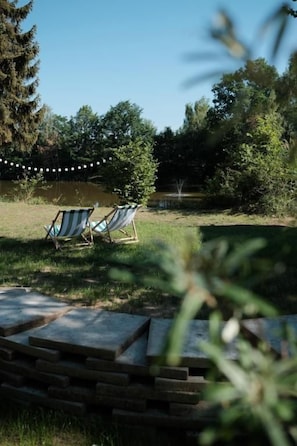 A grassy outdoor area with two striped lounge chairs and a stone pathway leading to a body of water. - kleiner Heidesee (Versmold)