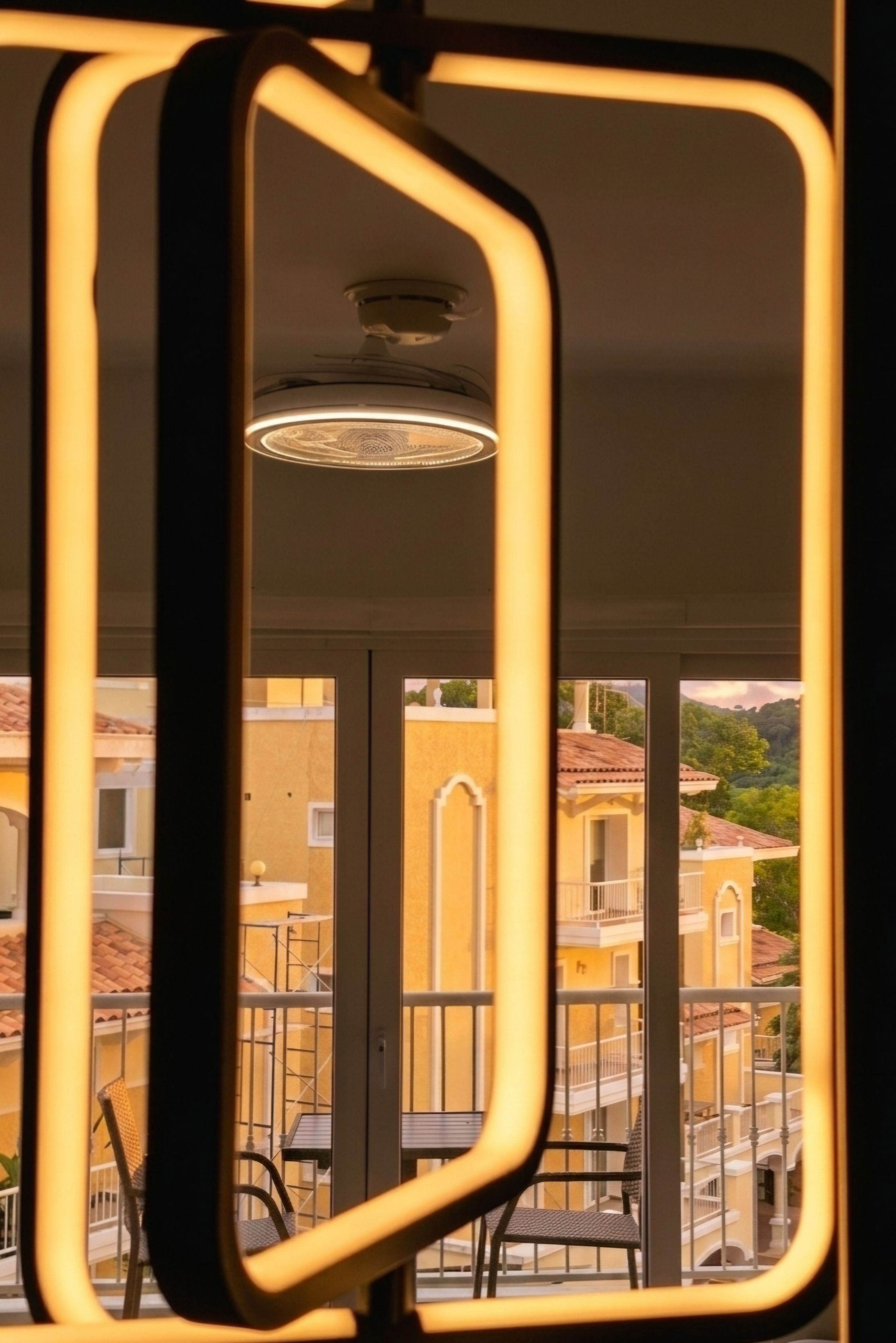 Une vue à travers une fenêtre ornée d’un cadre géométrique, dévoilant un balcon avec une balustrade et une chaise, ainsi que des bâtiments aux façades jaunes en arrière-plan.