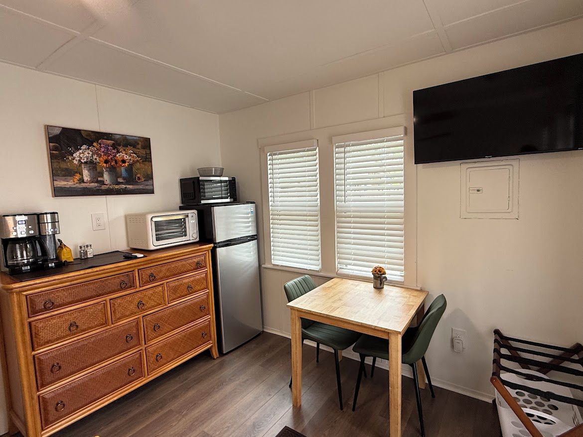 Une cuisine compacte équipée d’un four à micro-ondes, d’un réfrigérateur et d’une commode en bois. On y trouve une table à manger avec des chaises, ainsi qu’un tableau accroché au mur.