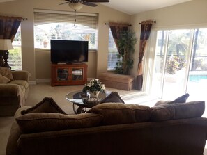 Ein Wohnzimmer mit einer Couch, einem Couchtisch mit einer Pflanze, einem Fernseher auf einem Schrank und einer Glasschiebetür, die zu einem Pool führt.