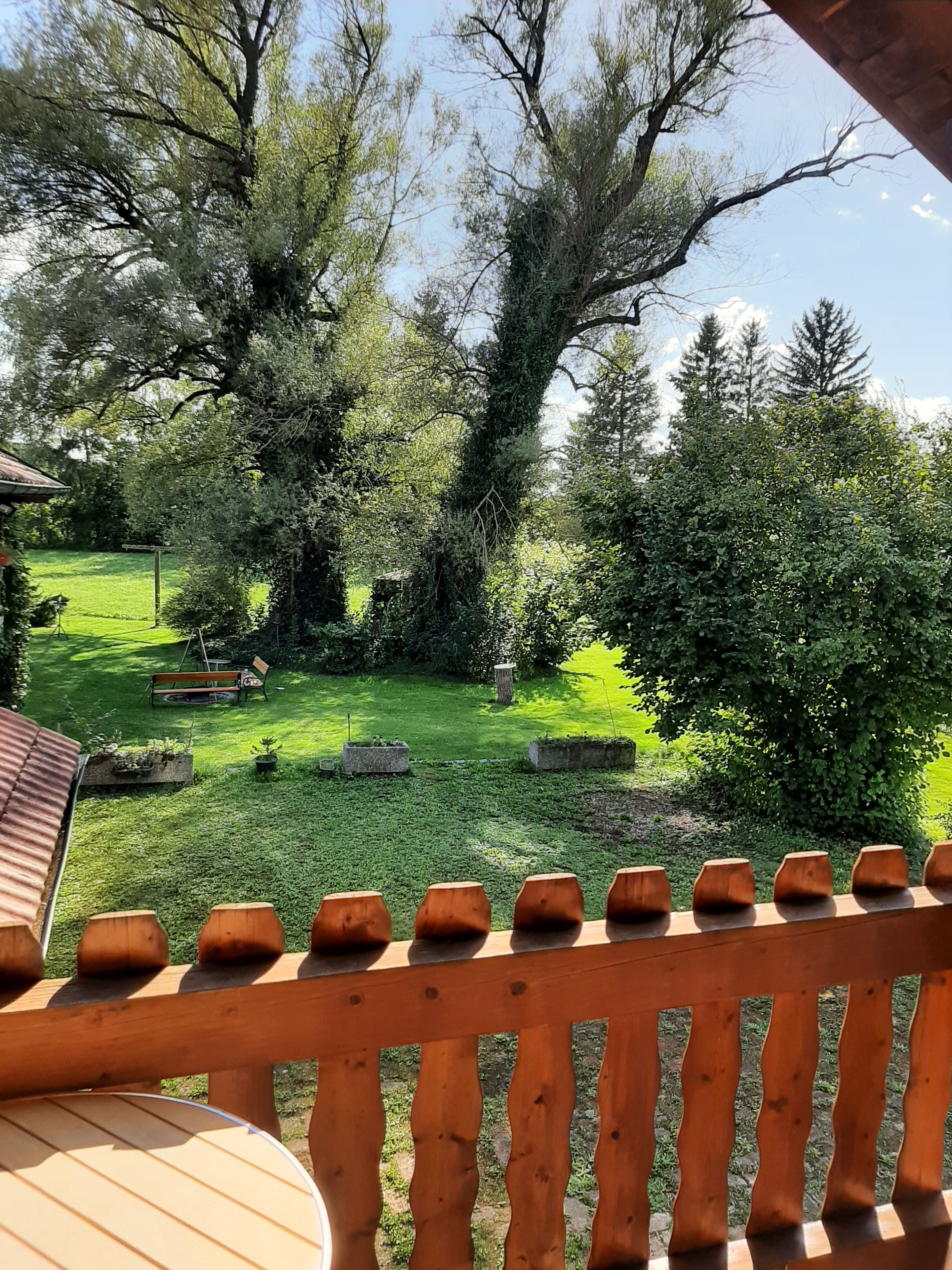 A wooden deck with a view of a well-maintained garden, including a lawn, trees, and a stone bench.