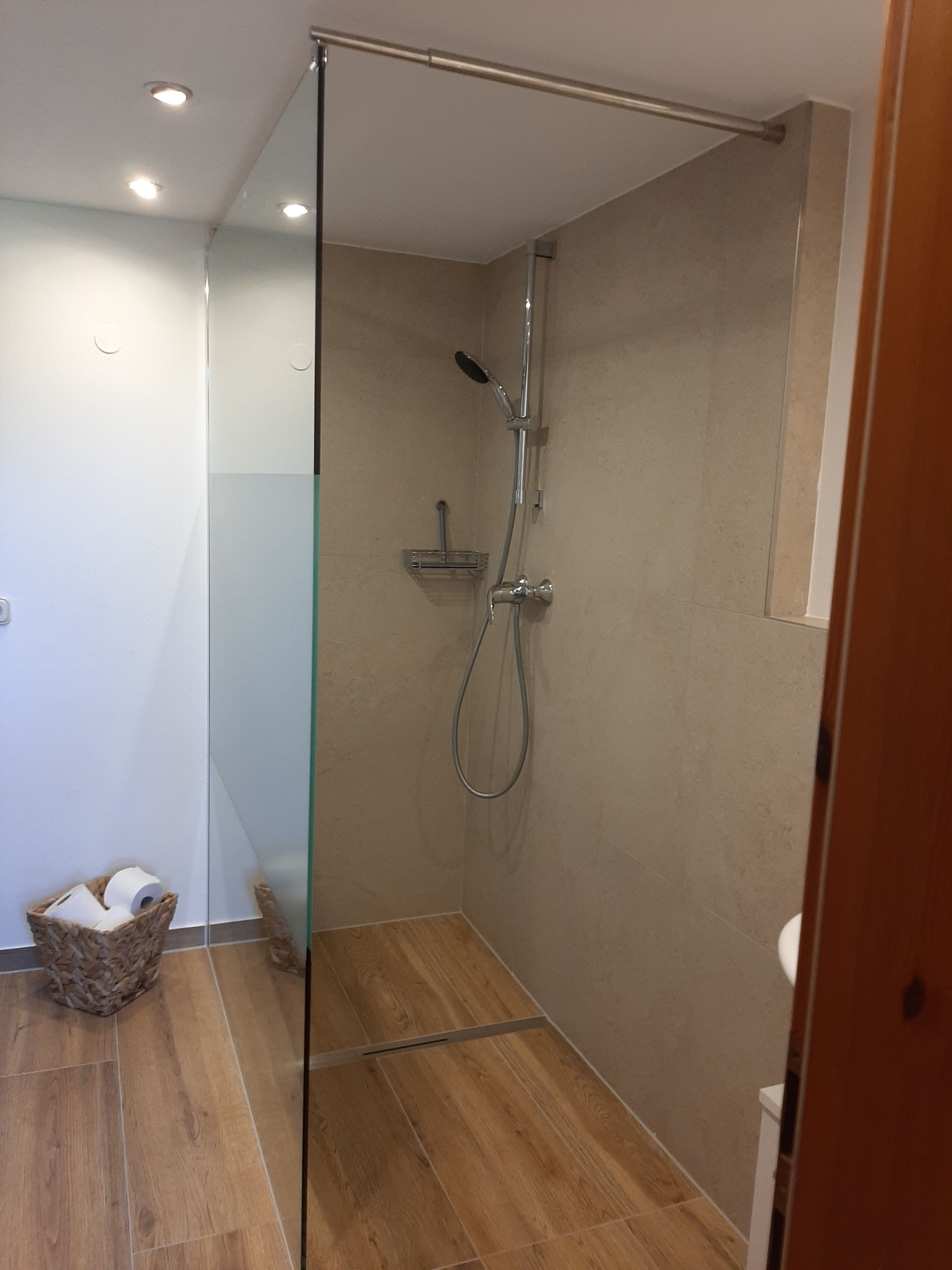 Ein modernes Badezimmer mit einer Duschkabine aus Glas, Holzboden und einer kleinen Mülltonne.