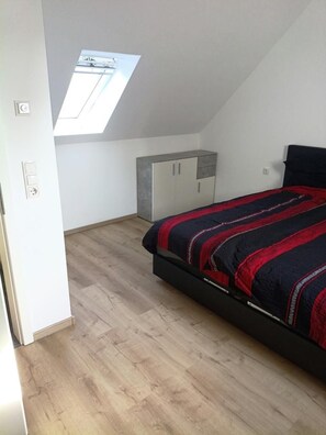A bedroom with a sloped ceiling, a skylight, a bed with a red and black bedspread, a wardrobe, and a light-colored floor. - New! Meerzeit Vacation Home (Nenndorf)