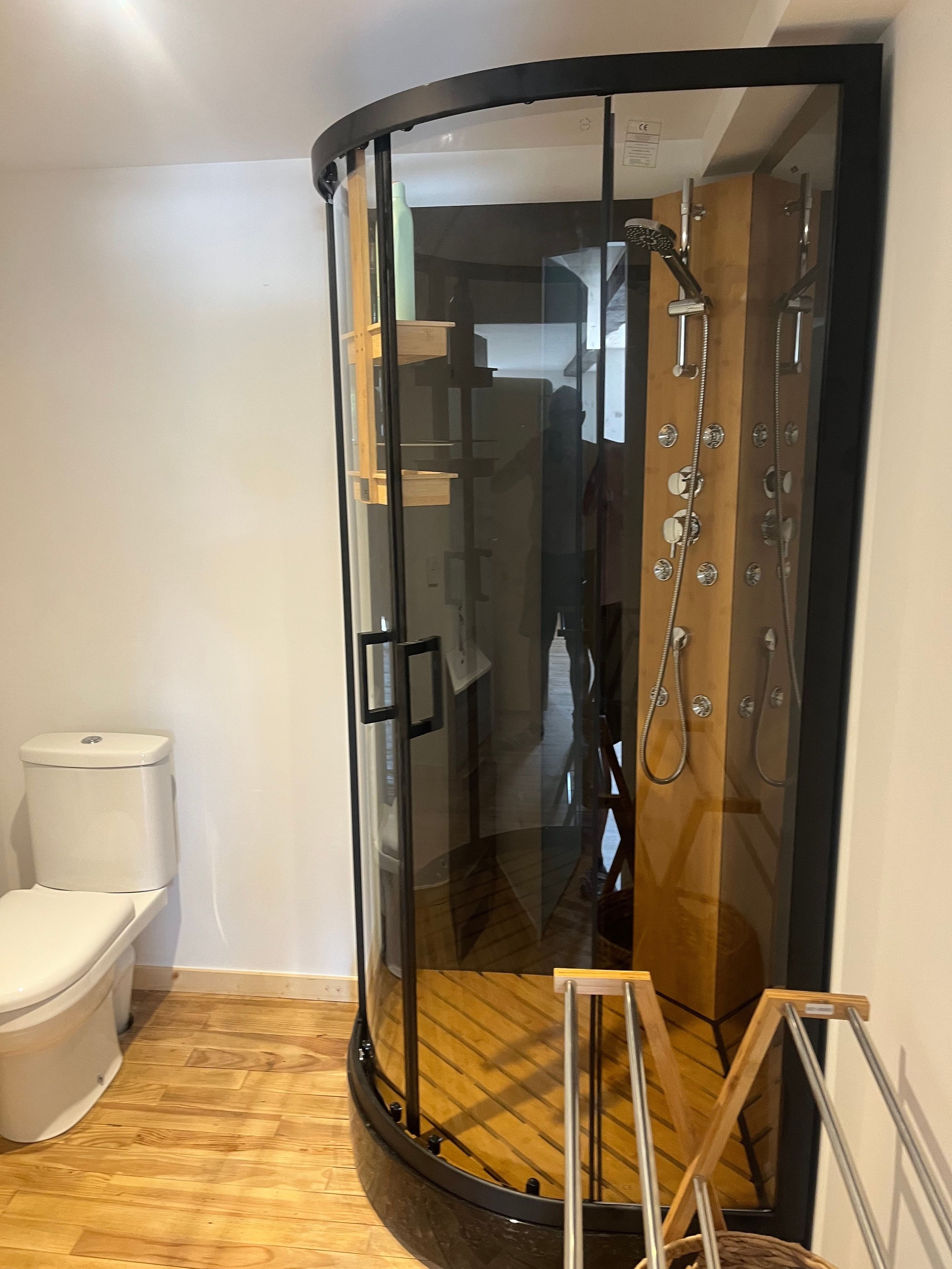 A glass-enclosed shower stall with a wooden floor, a toilet, and a small wooden stool.