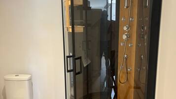 A glass-enclosed shower stall with a wooden floor, a toilet, and a small wooden stool.
