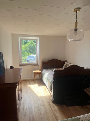 Ein Schlafzimmer mit einem hölzernen Bett, einem kleinen Nachttisch, einem Schreibtisch und einem Fenster mit Blick auf Grünflächen und ein Auto draußen.