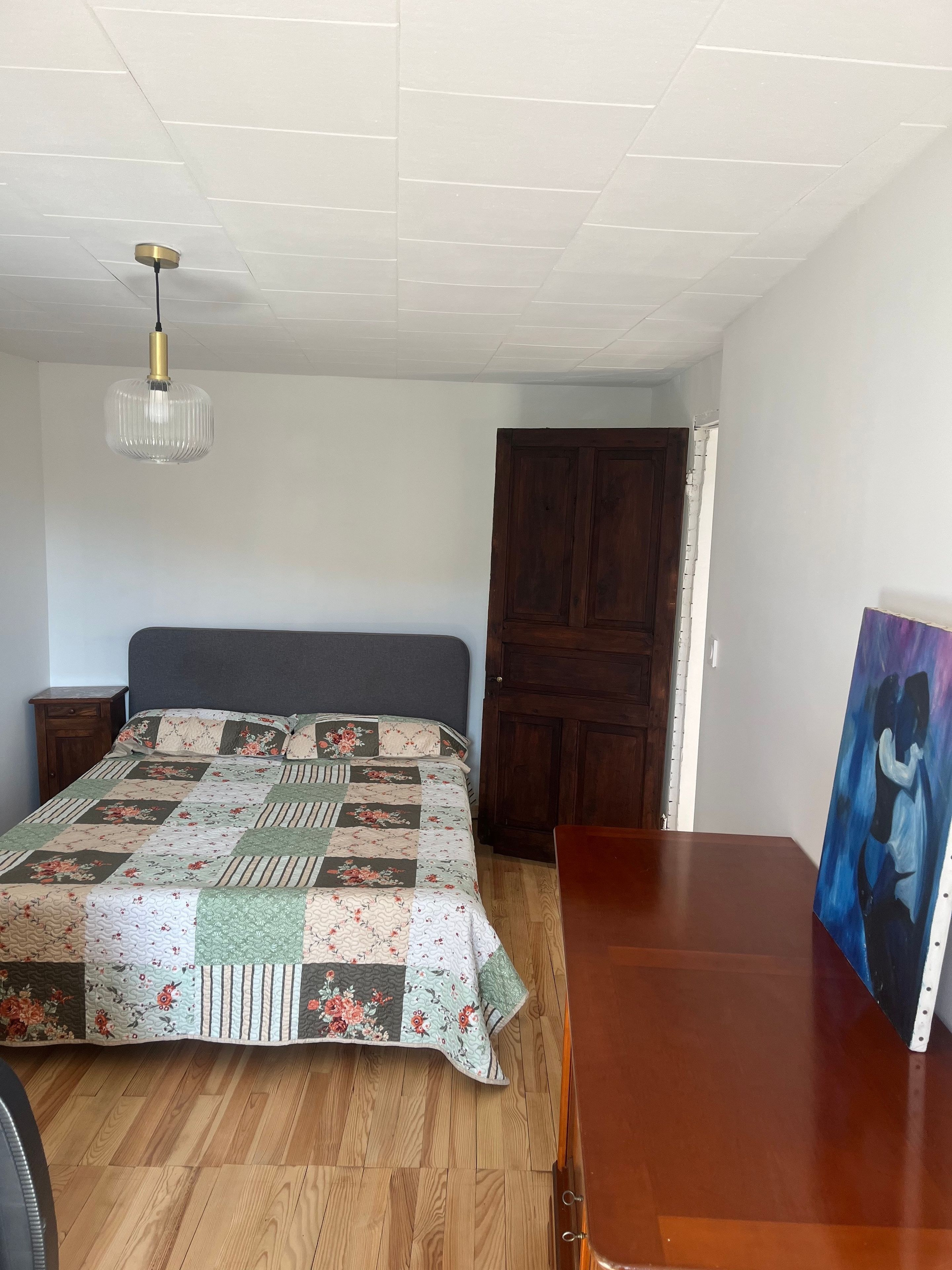 Un dormitori amb terres de fusta, un llit amb edredó floral, una tauleta de nit de fusta, una porta de fusta i un quadre a la paret.