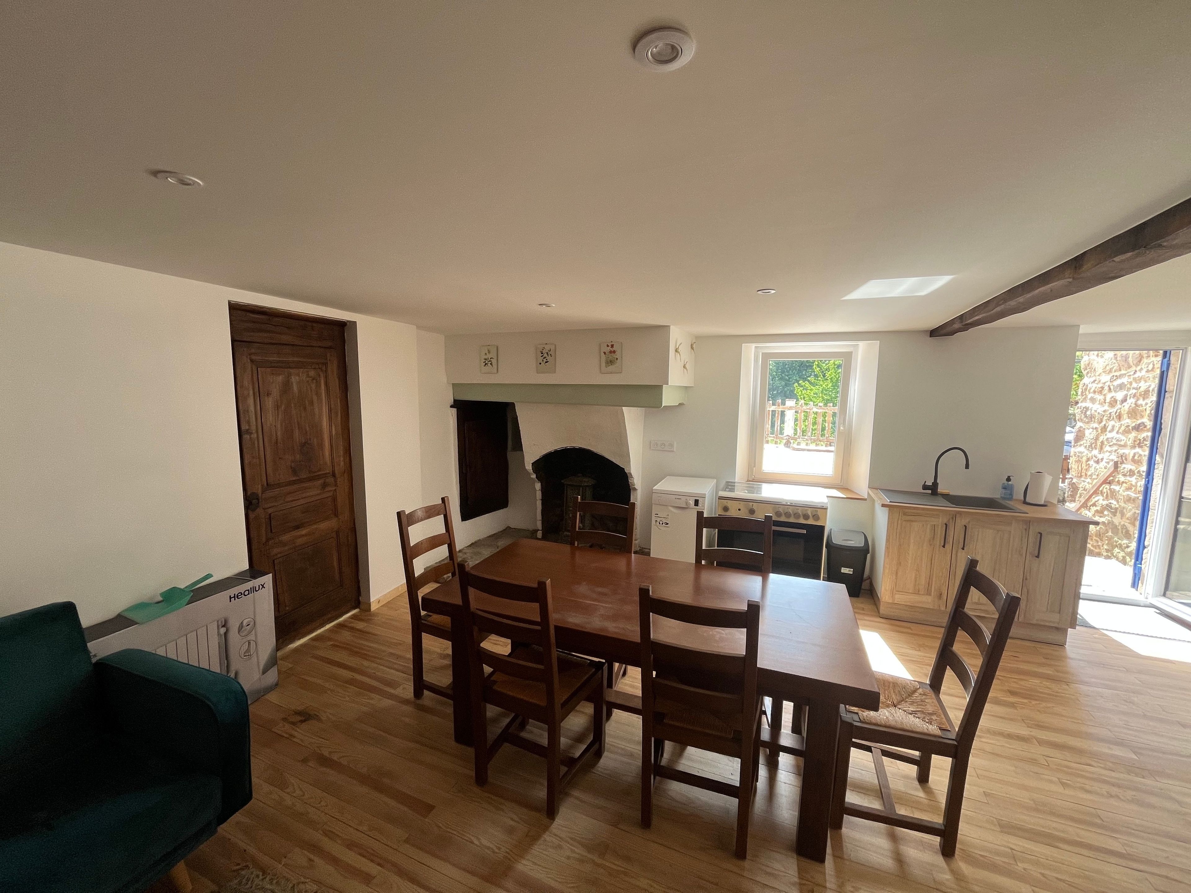A dining area with a wooden table and chairs, a kitchen with a window, and a stone wall outside.