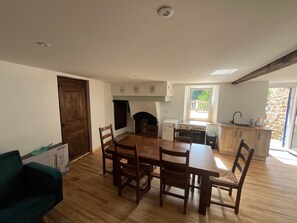 A dining area with a wooden table and chairs, a kitchen with a window, and a stone wall outside.