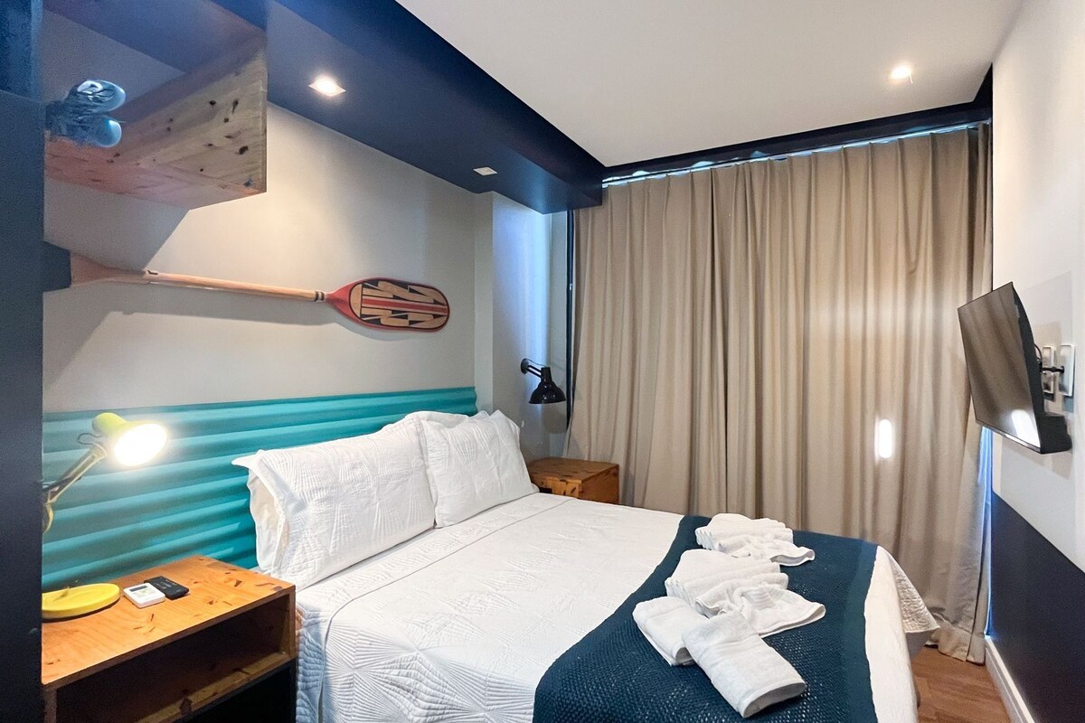 A hotel room with a bed, a desk with a lamp, a TV, and a large window with curtains.
