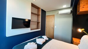 A hotel room with a bed, a flat-screen TV mounted on the wall, a wooden door, and an air conditioning unit. - Condominio Merepe Residence - Porto de Galinhas (Ipojuca)