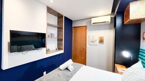 A hotel room with a bed, a TV mounted on the wall, a wooden door, and a small table with a towel on it. - Condominio Merepe Residence - Porto de Galinhas (Ipojuca)
