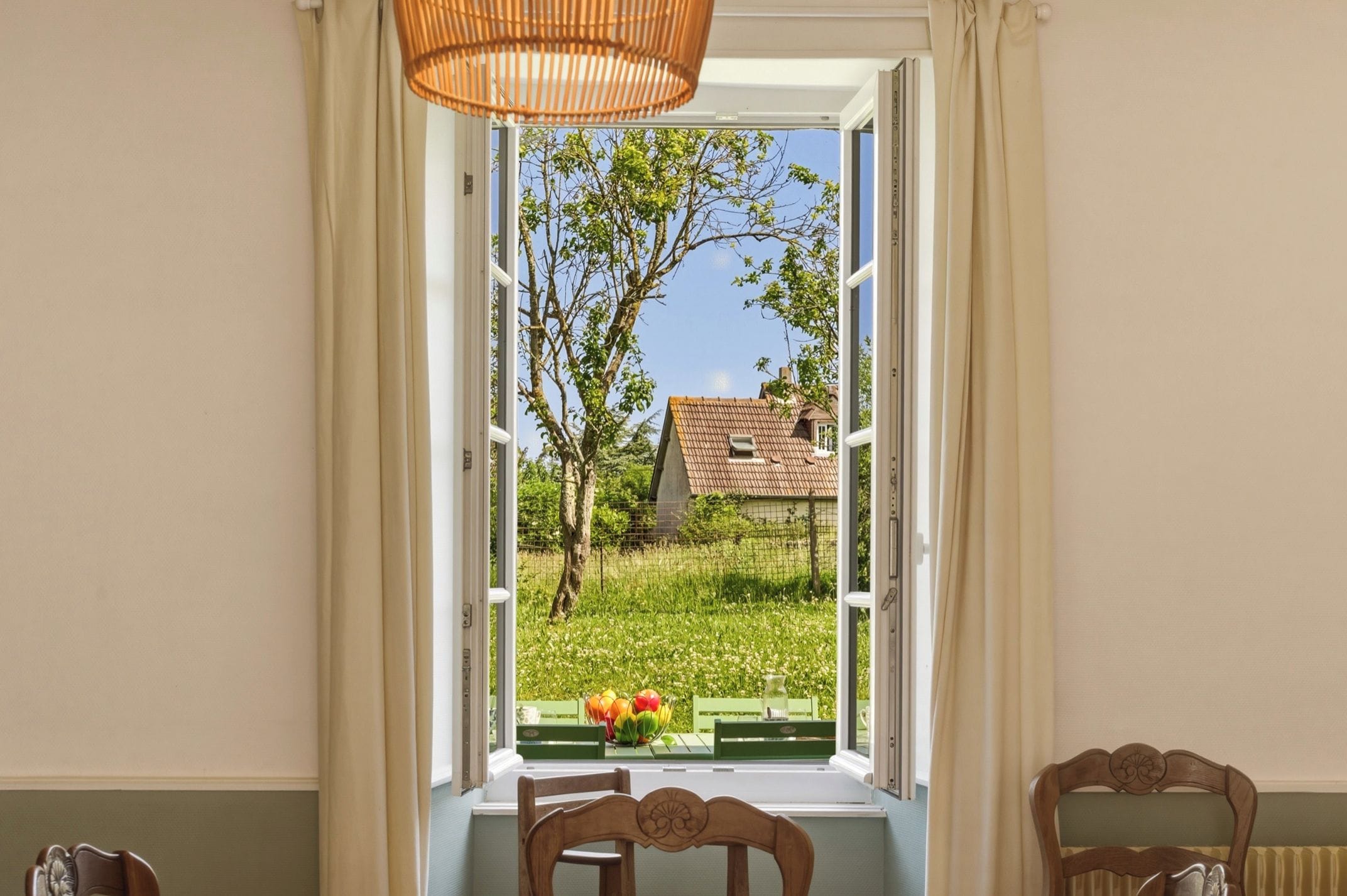 A room with a view through a window, a wicker chair, and a table with a bowl of fruit.