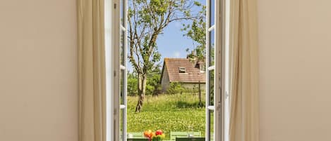 A room with a view through a window, a wicker chair, and a table with a bowl of fruit.