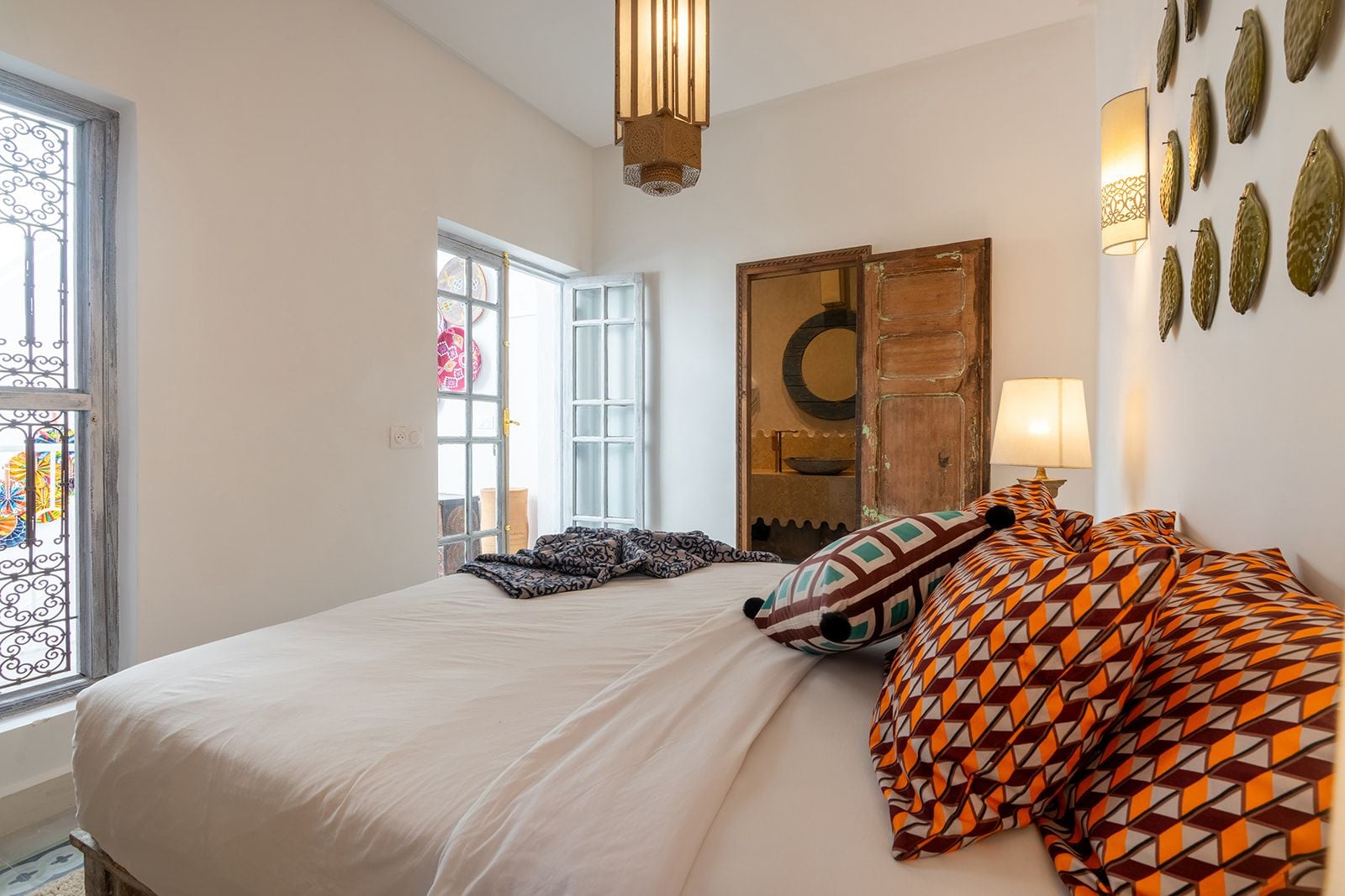 Une chambre avec un grand lit, une fenêtre ornée de barreaux en fer forgé, une armoire en bois et un luminaire fixé au mur.